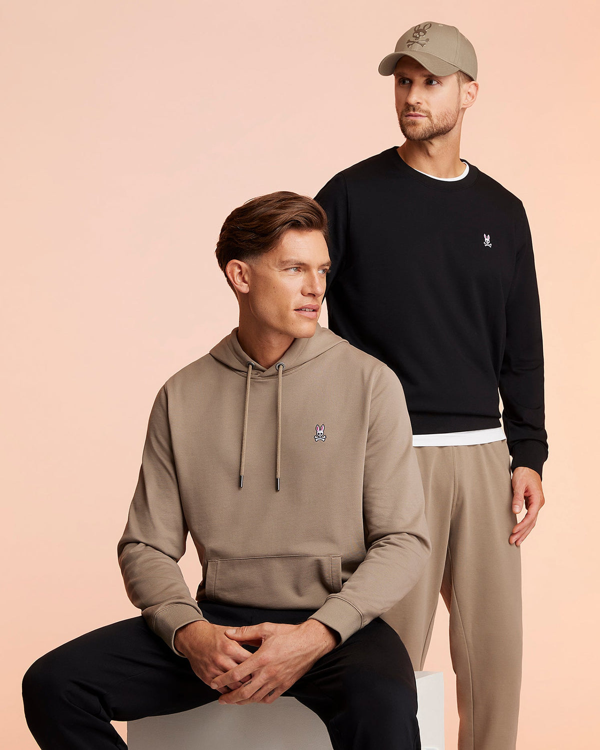 Two men sport casual Psycho Bunny attire with rabbit logos. One sits in a beige hoodie and black pants, while the other stands wearing a black sweatshirt, MENS FRENCH TERRY KNIT SWEATPANTS - B6P828ARFT in beige, and a matching cap. They pose against a peach background.