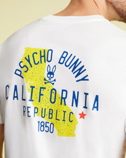 A person wearing the Psycho Bunny Men's California Tee (B6U552W1PC) made of breathable Pima cotton jersey, featuring a yellow graphic of California's state outline. The shirt prominently displays "PSYCHO BUNNY," "CALIFORNIA REPUBLIC," and "1850" in bold blue text, along with a small red star and the iconic bunny skull logo.