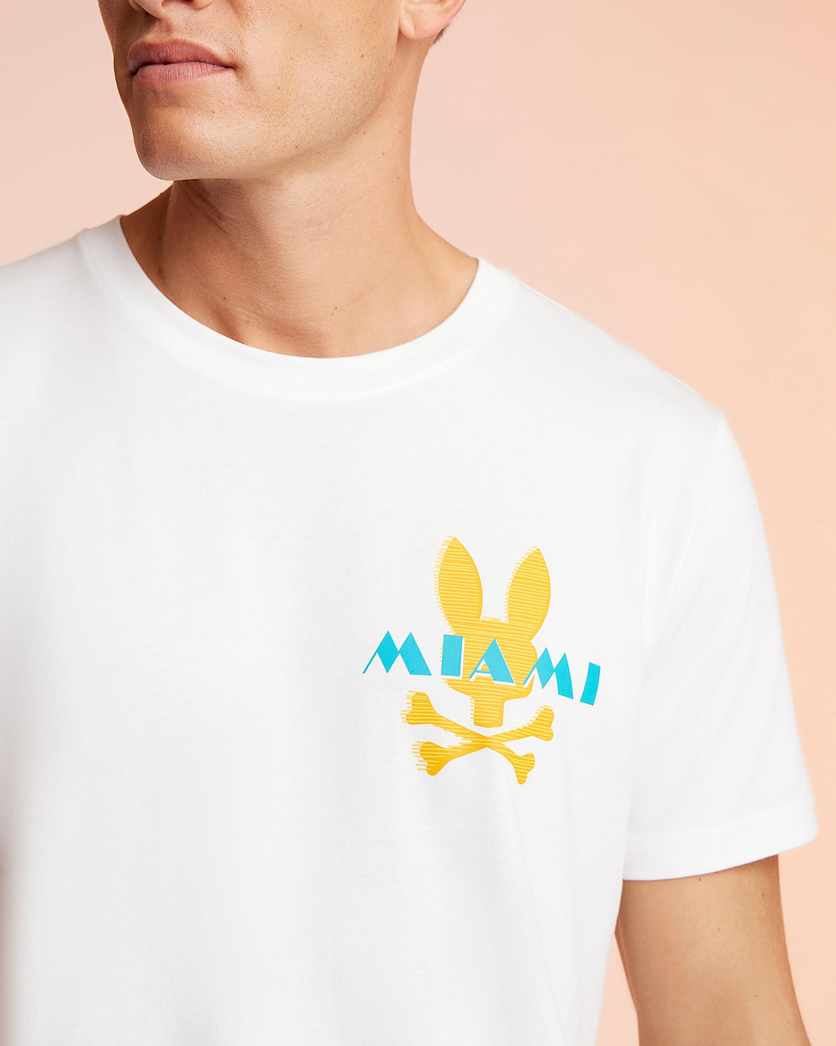 A person wearing a Psycho Bunny MENS MIAMI TEE - B6U550W1PC, a white T-shirt featuring a yellow bunny head and crossbones graphic above the word "MIAMI" in blue. The image focuses on the upper half of the Pima cotton T-shirt, showing part of the person's face and neck against a peach-colored background.