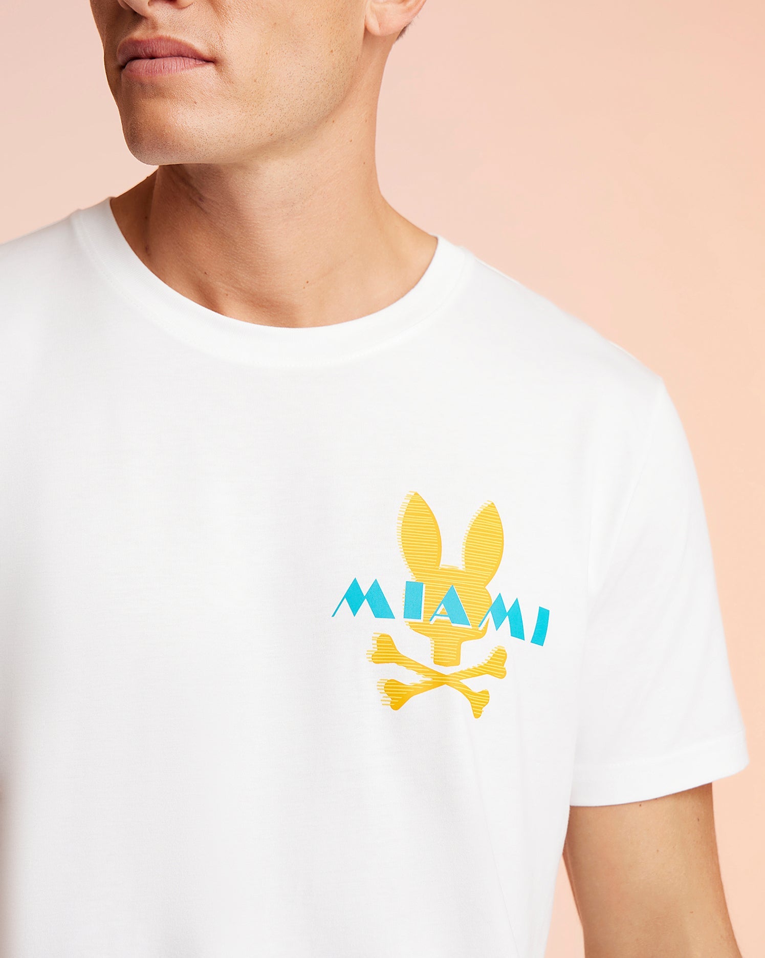 A person wearing a Psycho Bunny MENS MIAMI TEE - B6U550W1PC, a white T-shirt featuring a yellow bunny head and crossbones graphic above the word "MIAMI" in blue. The image focuses on the upper half of the Pima cotton T-shirt, showing part of the person's face and neck against a peach-colored background.
