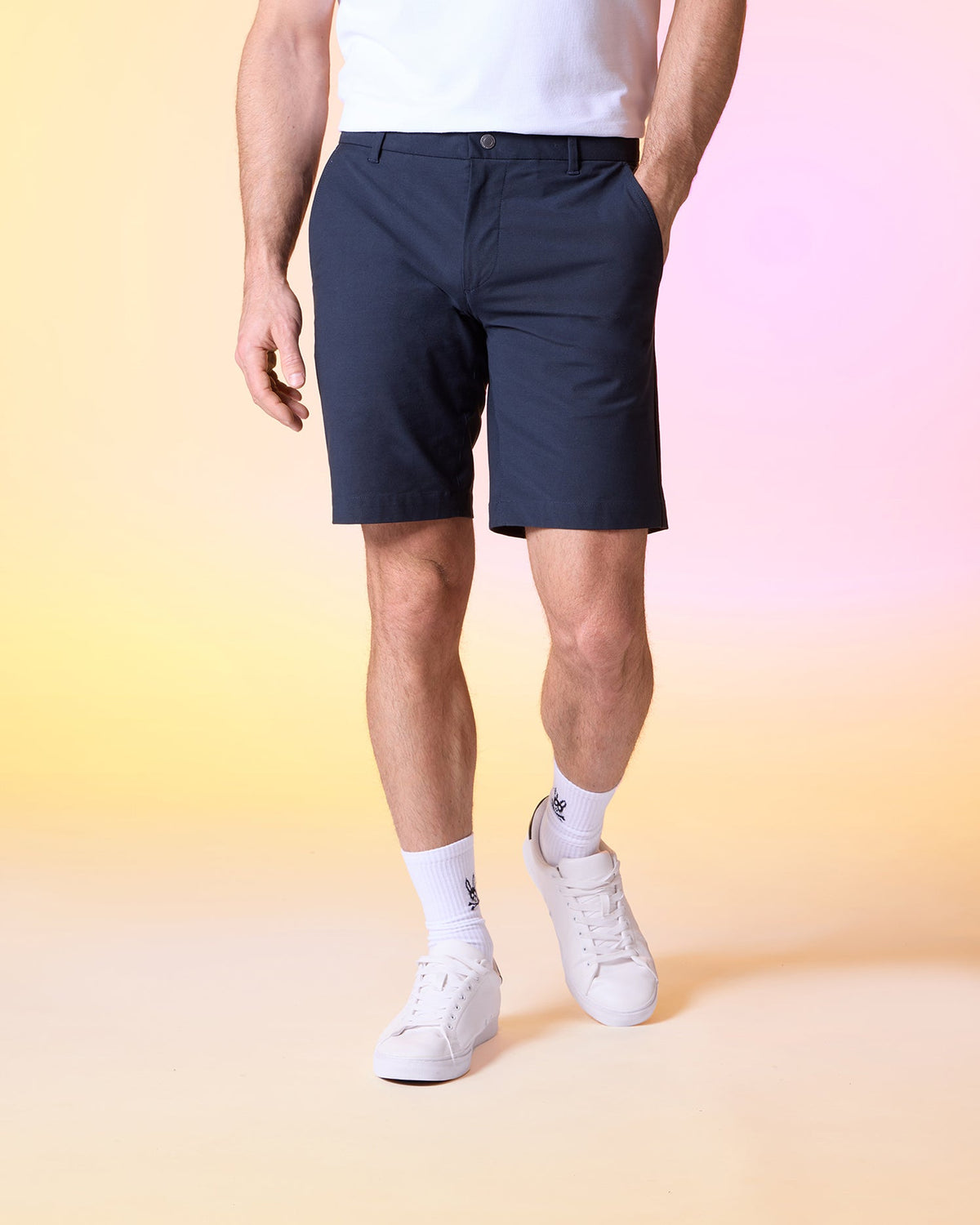 A person wearing a white t-shirt, Psycho Bunny MENS GABLE REGULAR FIT SPORT SHORT - B6R761ARCN made of durable stretch cotton-nylon, white socks, and white sneakers is standing against a pastel gradient background of yellow and pink. The person's hands are in their pockets, revealing a hidden zip pocket on the shorts, and their face is not visible.