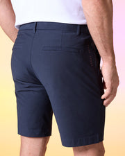 A man wearing a white shirt and navy blue Psycho Bunny MENS GABLE REGULAR FIT SPORT SHORT - B6R761ARCN stands with his back facing the camera. The durable stretch cotton-nylon shorts have belt loops, two back pockets with horizontal slits, a hidden zip pocket, and a small stitched design on the right side seam. The background is a gradient of yellow and pink.