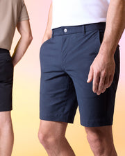 Two individuals are pictured from the waist down, showcasing casual attire. The person in the foreground is wearing a white top and navy blue MENS GABLE REGULAR FIT SPORT SHORT - B6R761ARCN by Psycho Bunny with hidden zip pockets for convenience. The person in the background sports a tan shirt and black shorts. The backdrop features soft pink and yellow hues.