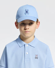 A young boy is wearing the Psycho Bunny KIDS NEW CASTLE EMBROIDERED BASEBALL CAP, featuring a rabbit skull logo and an adjustable back strap for a comfortable fit. He wears a light blue polo shirt and looks directly at the camera against a plain background.