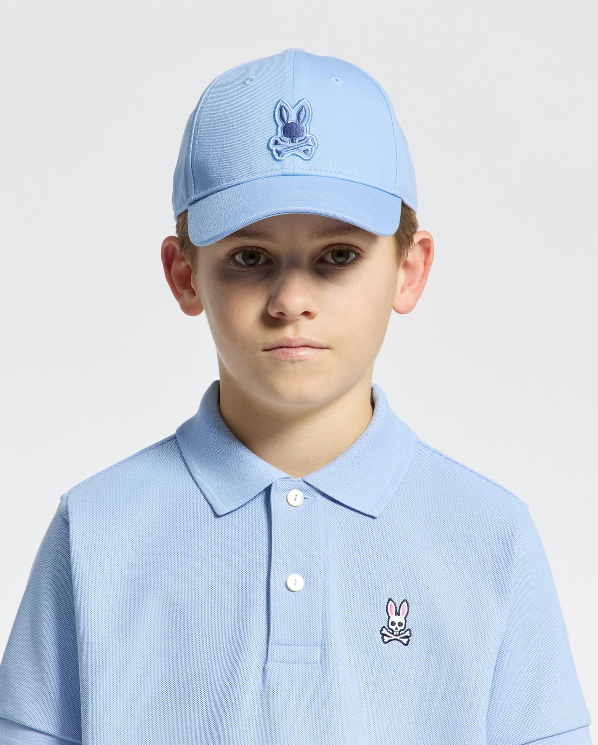A young boy is wearing the Psycho Bunny KIDS NEW CASTLE EMBROIDERED BASEBALL CAP, featuring a rabbit skull logo and an adjustable back strap for a comfortable fit. He wears a light blue polo shirt and looks directly at the camera against a plain background.