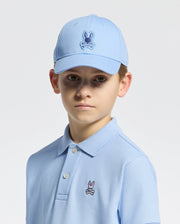 A young boy wears the Psycho Bunny KIDS NEW CASTLE EMBROIDERED BASEBALL CAP - B0A547F200 with an adjustable back strap and a matching light blue polo, both 100% cotton and featuring a cartoon rabbit logo. He stands against a white background, gazing directly at the camera with a neutral expression.