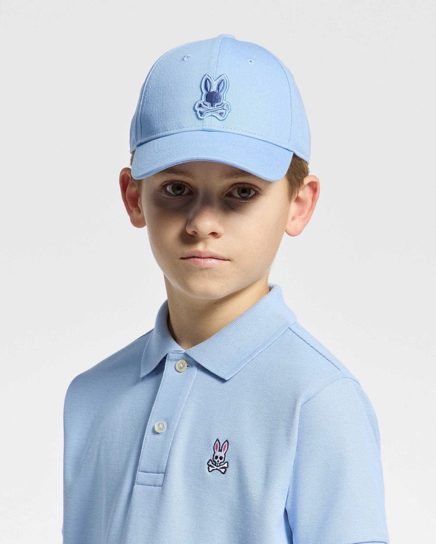 A young boy wears the Psycho Bunny KIDS NEW CASTLE EMBROIDERED BASEBALL CAP - B0A547F200 with an adjustable back strap and a matching light blue polo, both 100% cotton and featuring a cartoon rabbit logo. He stands against a white background, gazing directly at the camera with a neutral expression.