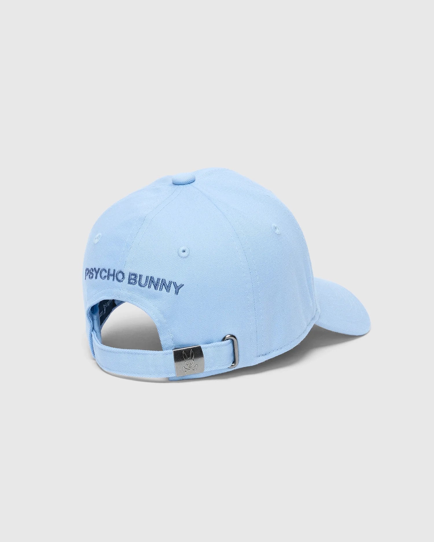 The KIDS NEW CASTLE EMBROIDERED BASEBALL CAP - B0A547F200 by Psycho Bunny is a light blue, 100% cotton cap with "PSYCHO BUNNY" embroidered in black on the back. It includes an adjustable strap with a metal buckle and small logo, set against a plain light gray background.