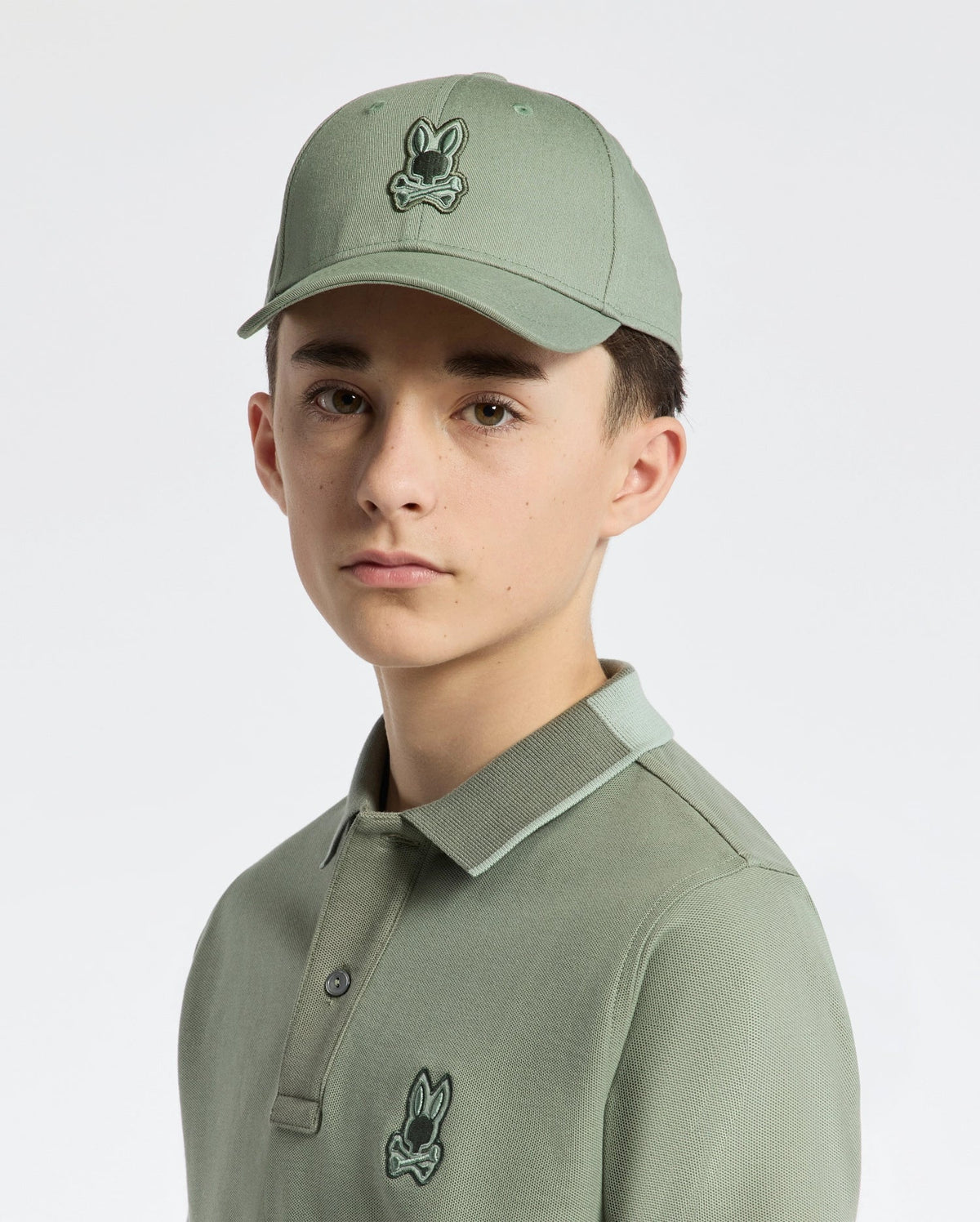 Against a white background, a young person wears a Psycho Bunny KIDS NEW CASTLE BASEBALL CAP in sage green with an adjustable back strap and a polo shirt showcasing an embroidered bunny logo.