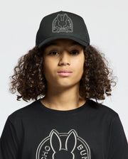 A person with curly hair wears a black Psycho Bunny hat named "Kids Melbourne Graphic Trucker Hat - B0A548F200," along with a matching black shirt. Each piece features an embroidered bunny logo and snap-back straps, against a plain background, while the person faces forward.