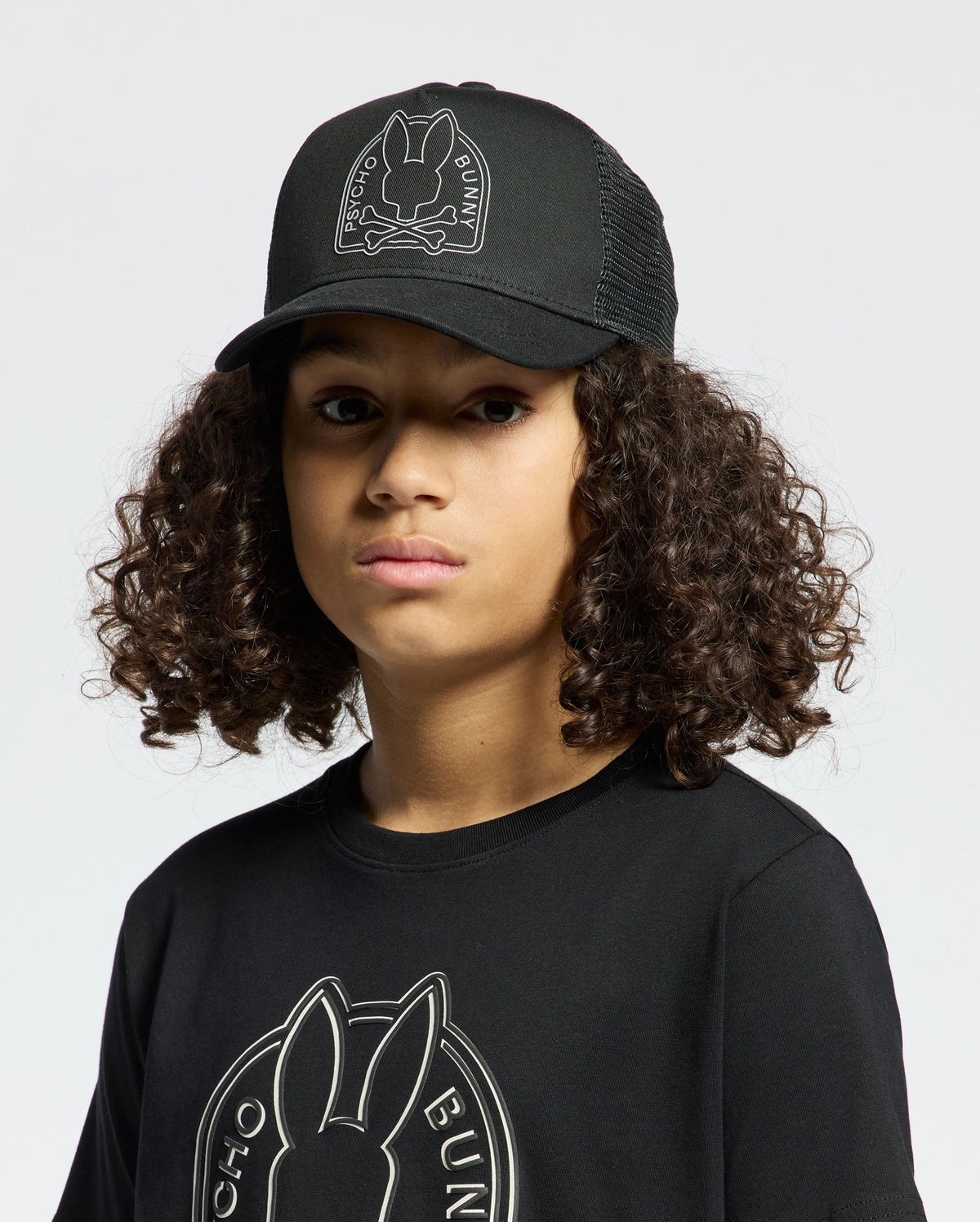 A person with curly hair wears a Psycho Bunny KIDS MELBOURNE GRAPHIC TRUCKER HAT and T-shirt, both with an embroidered bunny icon, against a plain white backdrop.