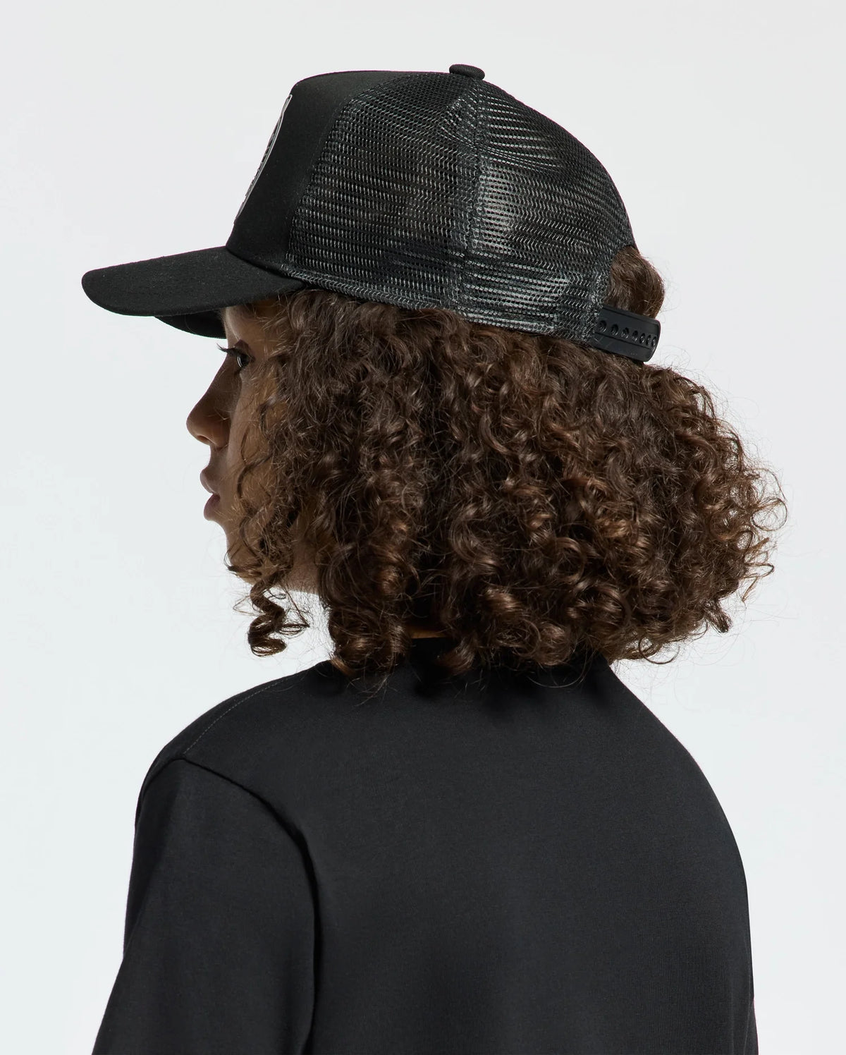 A person with curly brown hair is wearing a black mesh KIDS MELBOURNE GRAPHIC TRUCKER HAT by Psycho Bunny featuring an embroidered icon and a black shirt, gazing to the side against a neutral backdrop.