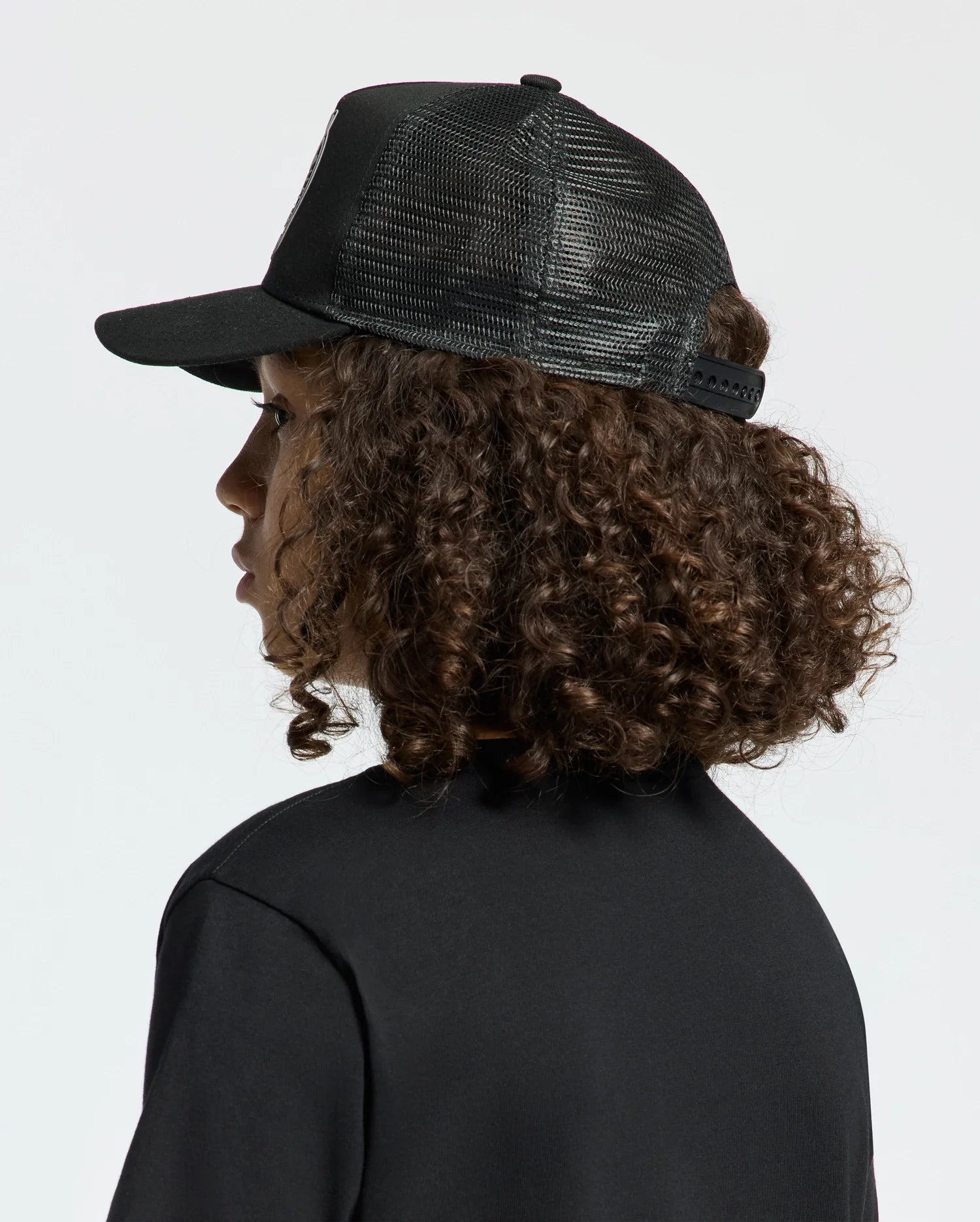 A person with curly brown hair is wearing a black mesh KIDS MELBOURNE GRAPHIC TRUCKER HAT by Psycho Bunny featuring an embroidered icon and a black shirt, gazing to the side against a neutral backdrop.