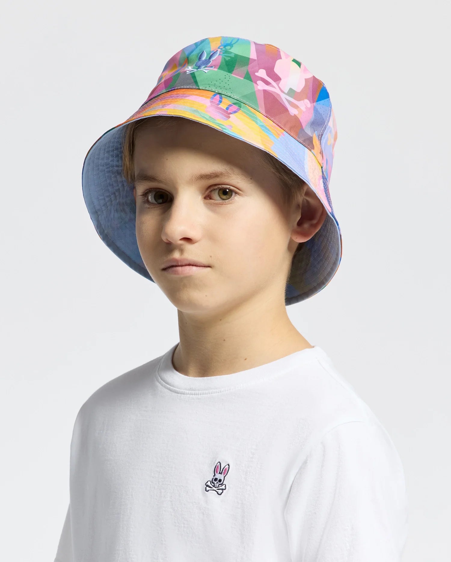 A young person wearing the KIDS MILOS BUCKET REVERSIBLE HAT - B0A550F200 and a plain white shirt with an embroidered Psycho Bunny logo stands against a plain background, looking into the camera.
