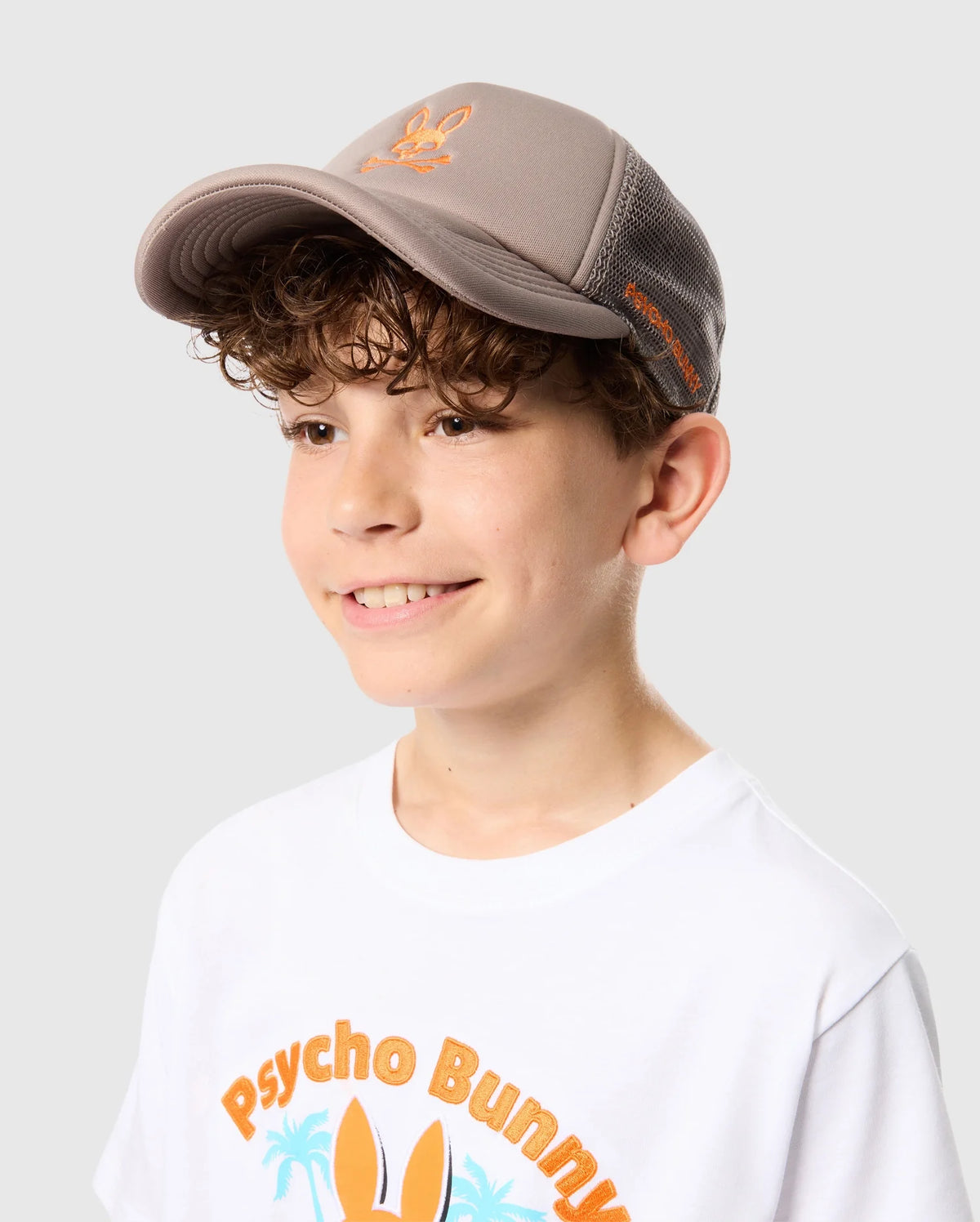 A smiling boy wears a light brown Psycho Bunny Kids Paris Trucker Cap (B0A591C200) with mesh panels, paired with a white t-shirt featuring "Psycho Bunny" in vibrant letters. The cap has an orange bunny logo with crossed bones, set against a light gray backdrop.