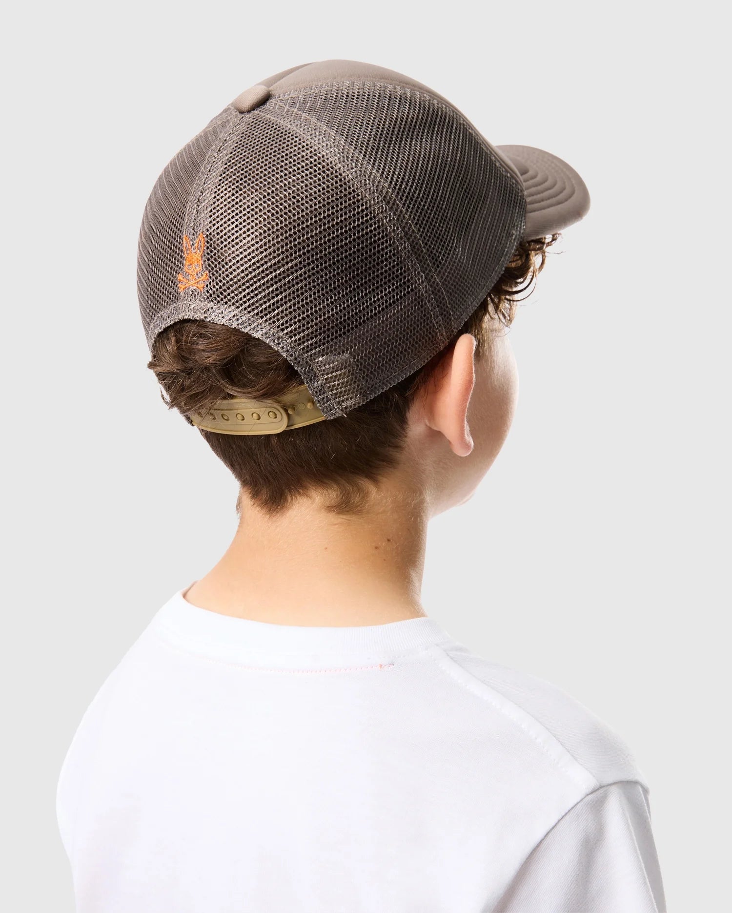 A person is seen from behind wearing a Psycho Bunny KIDS PARIS TRUCKER CAP - B0A591C200, featuring gray mesh paneling, an adjustable strap, and a small orange Bunny logo. The look is complemented by a white T-shirt.