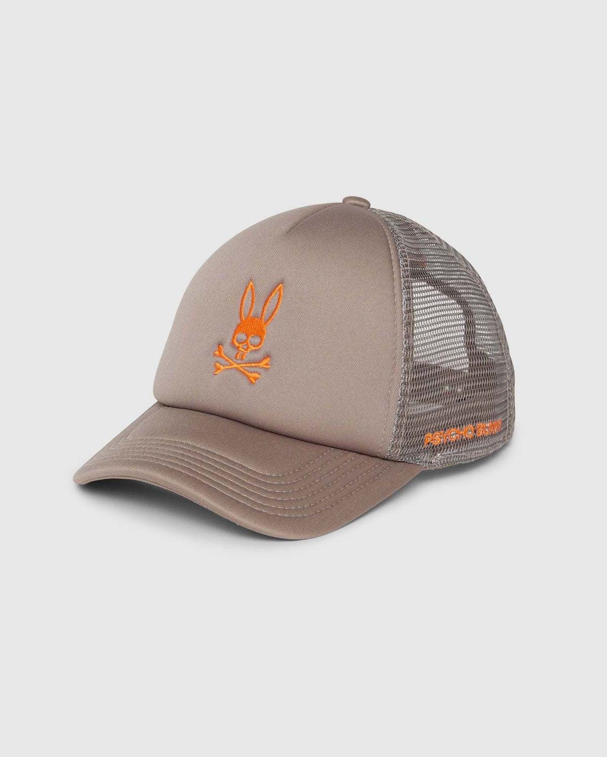 The KIDS PARIS TRUCKER CAP (B0A591C200) by Psycho Bunny features a beige hue with a mesh back and an orange Bunny logo of a rabbit's head above crossbones. The brim has visible stitching, and the side is embroidered with text. This cap blends playful design with trendy flair.