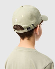 A person with short, curly hair is seen from behind, wearing a light green cotton twill KIDS KAYDEN BASEBALL CAP - B0A677C200 and a matching t-shirt. The cap, from Psycho Bunny, has an adjustable strap with a metal clasp above the nape of the neck and an embroidered white Bunny logo above the strap. The background is plain white.