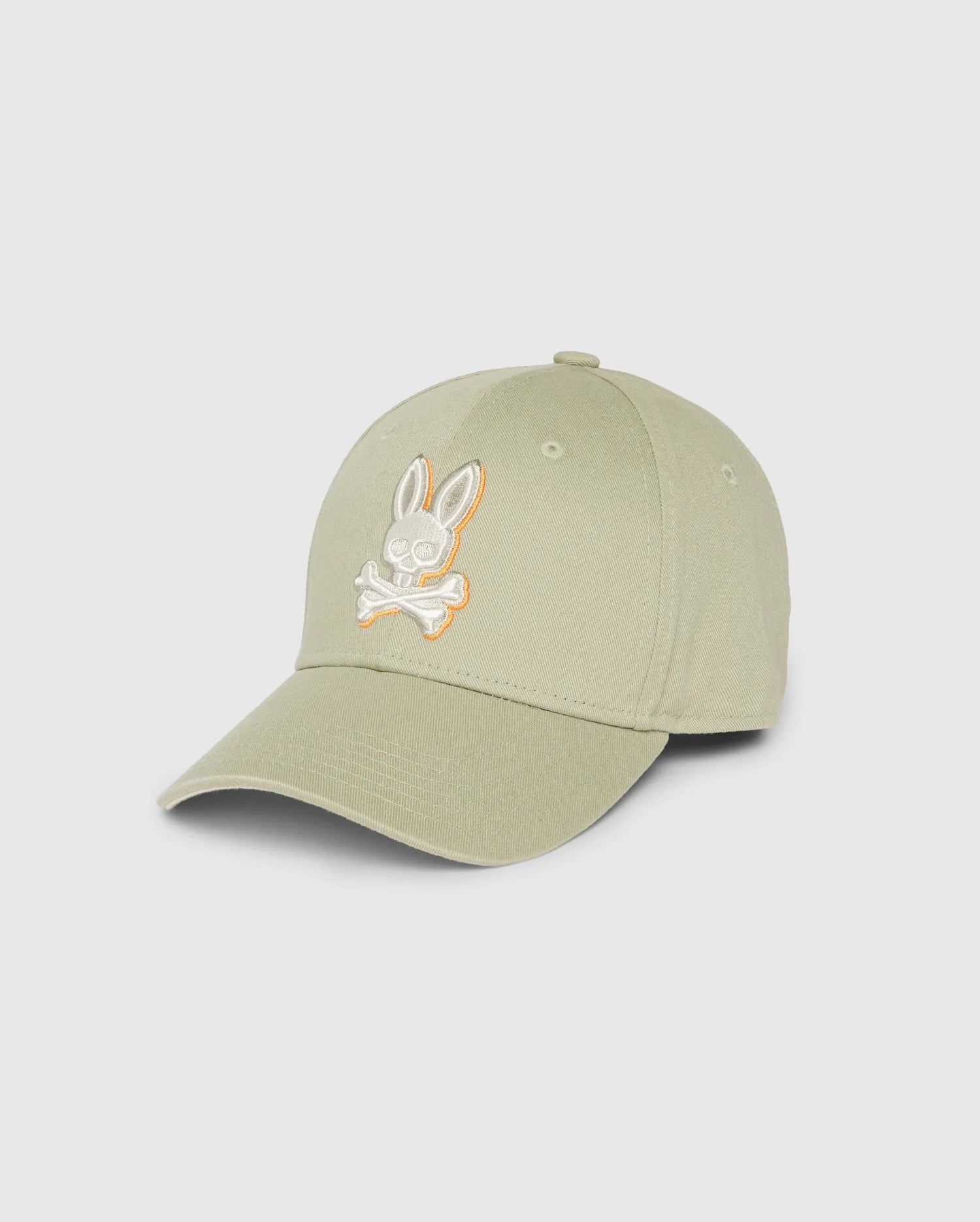 Introducing the KIDS KAYDEN BASEBALL CAP - B0A677C200 by Psycho Bunny: a light olive green cotton twill cap, adorned with a striking embroidered Bunny logo featuring crossed bones in white and orange on the front. This cap is designed with a curved brim and six panels, each with ventilation eyelets for added comfort.