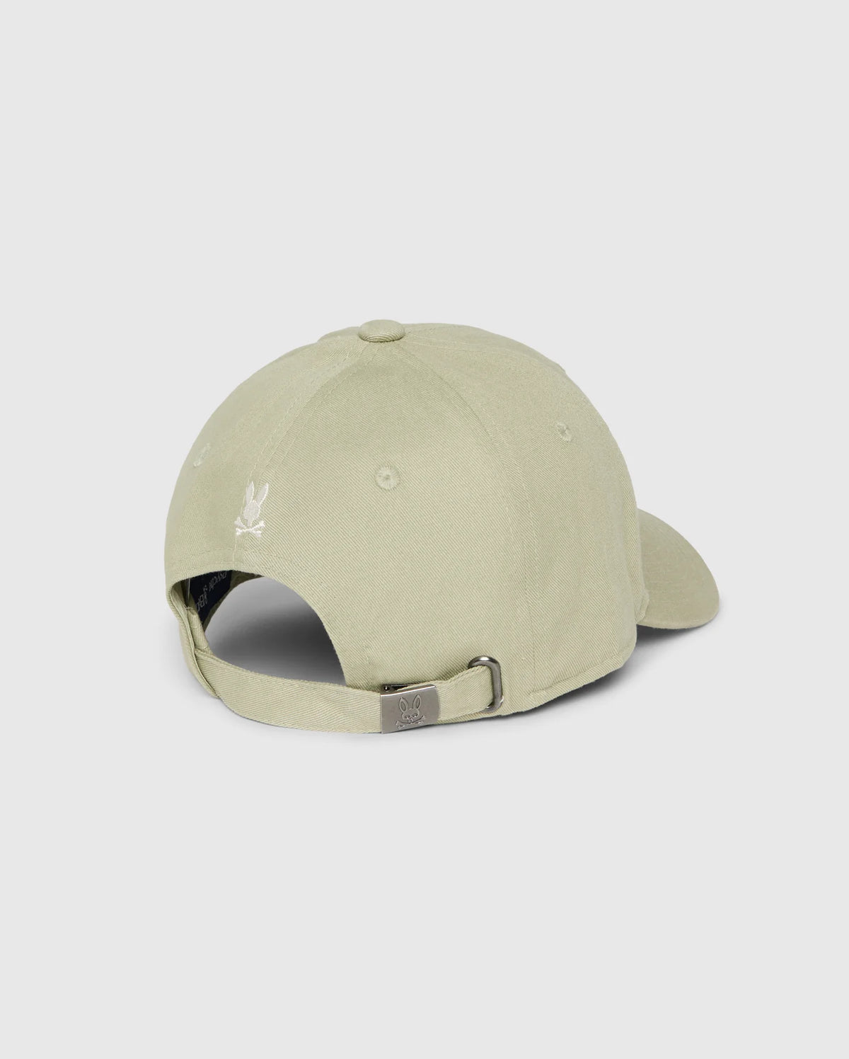 A light beige cotton twill baseball cap from Psycho Bunny, named the Kids Kayden Baseball Cap - B0A677C200, is viewed from the back. The cap features a metal buckle for size adjustment and an embroidered Bunny logo above the opening. The fabric has a smooth texture, and the cap is displayed against a plain, neutral background.