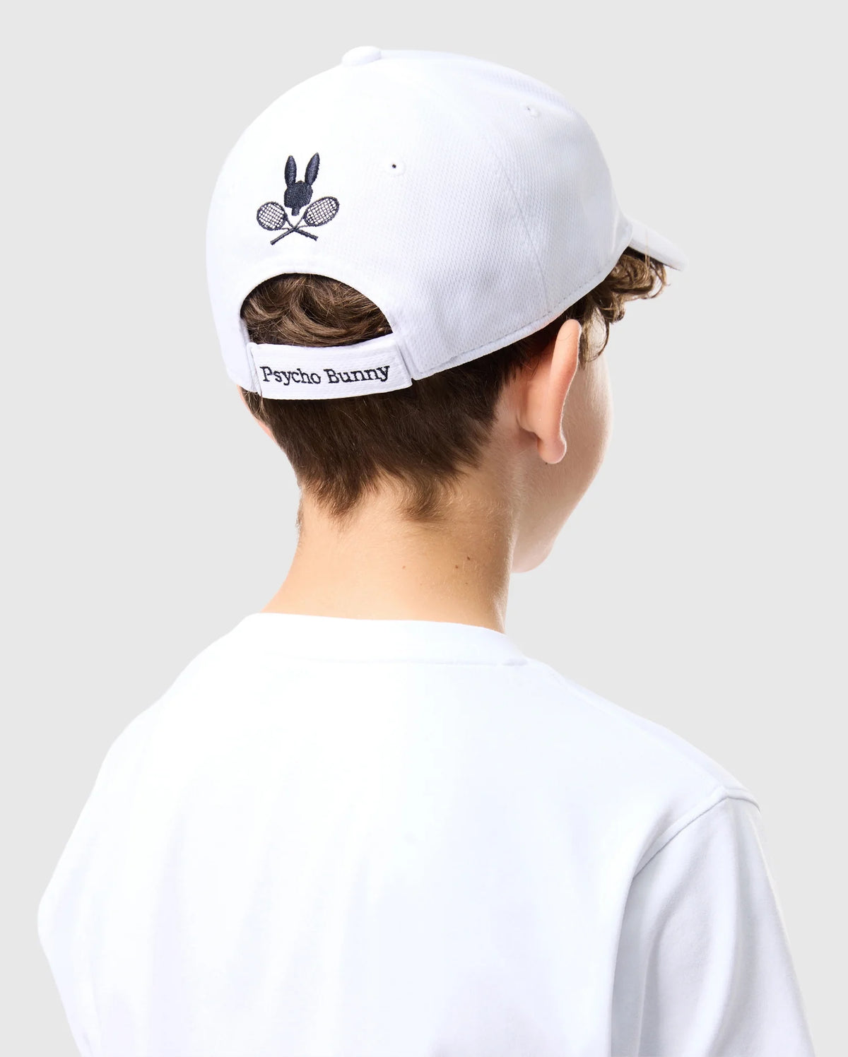 A person wears the Kids Courtside Sport Cap (B0A685C200) showcasing the tennis-inspired Bunny logo with crossed racquets, and "Psycho Bunny" text on the back strap. They pair it with a white shirt against a light gray background for a classic accessory look.