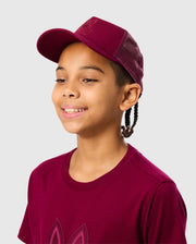 A young person smiles wearing a maroon KIDS MALTON TRUCKER CAP (B0A797D200) and matching T-shirt by Psycho Bunny, featuring mesh detailing on the back, set against a light gray background.