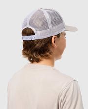 A person with medium-length hair is wearing a Psycho Bunny KIDS MALTON TRUCKER CAP - B0A797D200 and a light beige cotton t-shirt. The person is facing away from the camera, showcasing the back of the trucker cap and the upper part of the t-shirt against a plain light grey background.