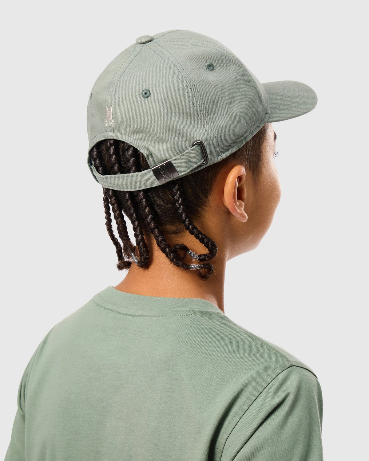 A person in a sporty, sage green KIDS ZACK BASEBALL CAP - B0A886D200 from Psycho Bunny and a matching t-shirt is shown from behind. The woven cotton cap, featuring a small embroidered logo, complements their braided hair against a plain light gray background.
