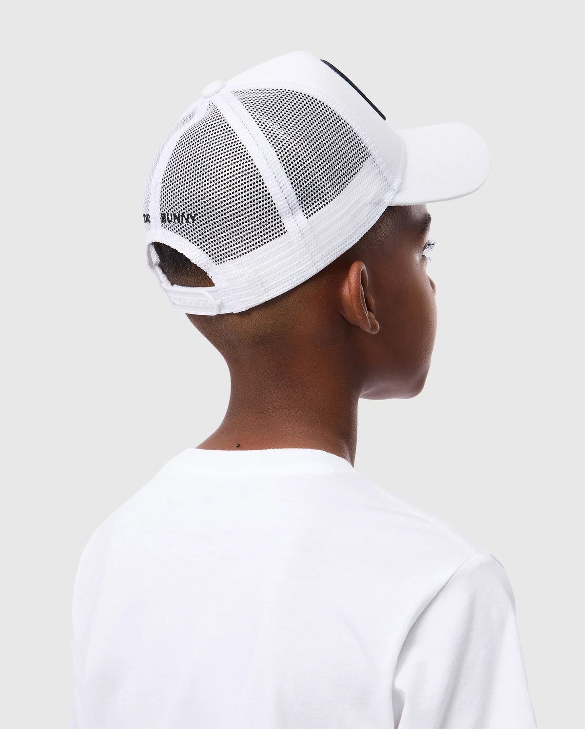 A person sporting a white mesh baseball cap with "BAD BUNNY" on the back is seen from behind. The cap resembles the KIDS BUNNY PATCH TRUCKER CAP - B0A965C200 by Psycho Bunny, featuring a solid white front and mesh back. They also wear a plain white t-shirt against a light gray backdrop.