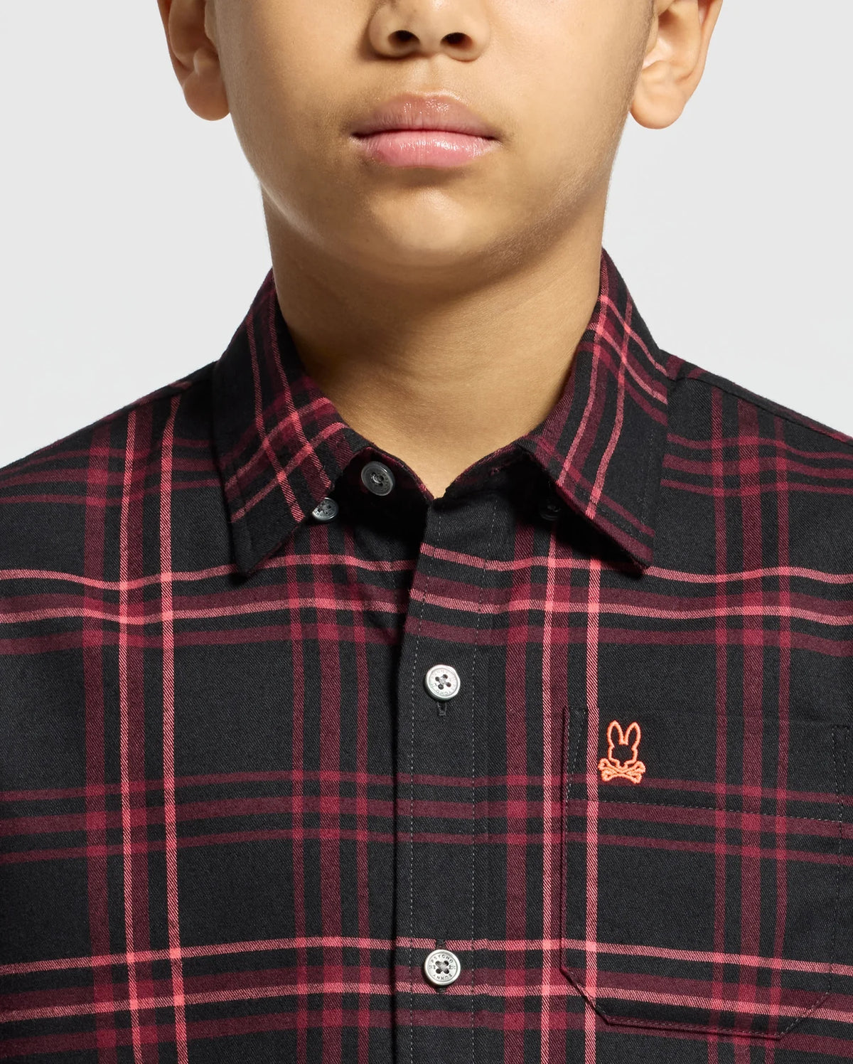 A person is wearing a Psycho Bunny KIDS HAYES PLAID SHIRT in black and red plaid Pima cotton, featuring a pink embroidered rabbit logo on the chest. The image highlights the collar and buttons on the upper torso.