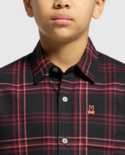 A person is wearing a Psycho Bunny KIDS HAYES PLAID SHIRT in black and red plaid Pima cotton, featuring a pink embroidered rabbit logo on the chest. The image highlights the collar and buttons on the upper torso.