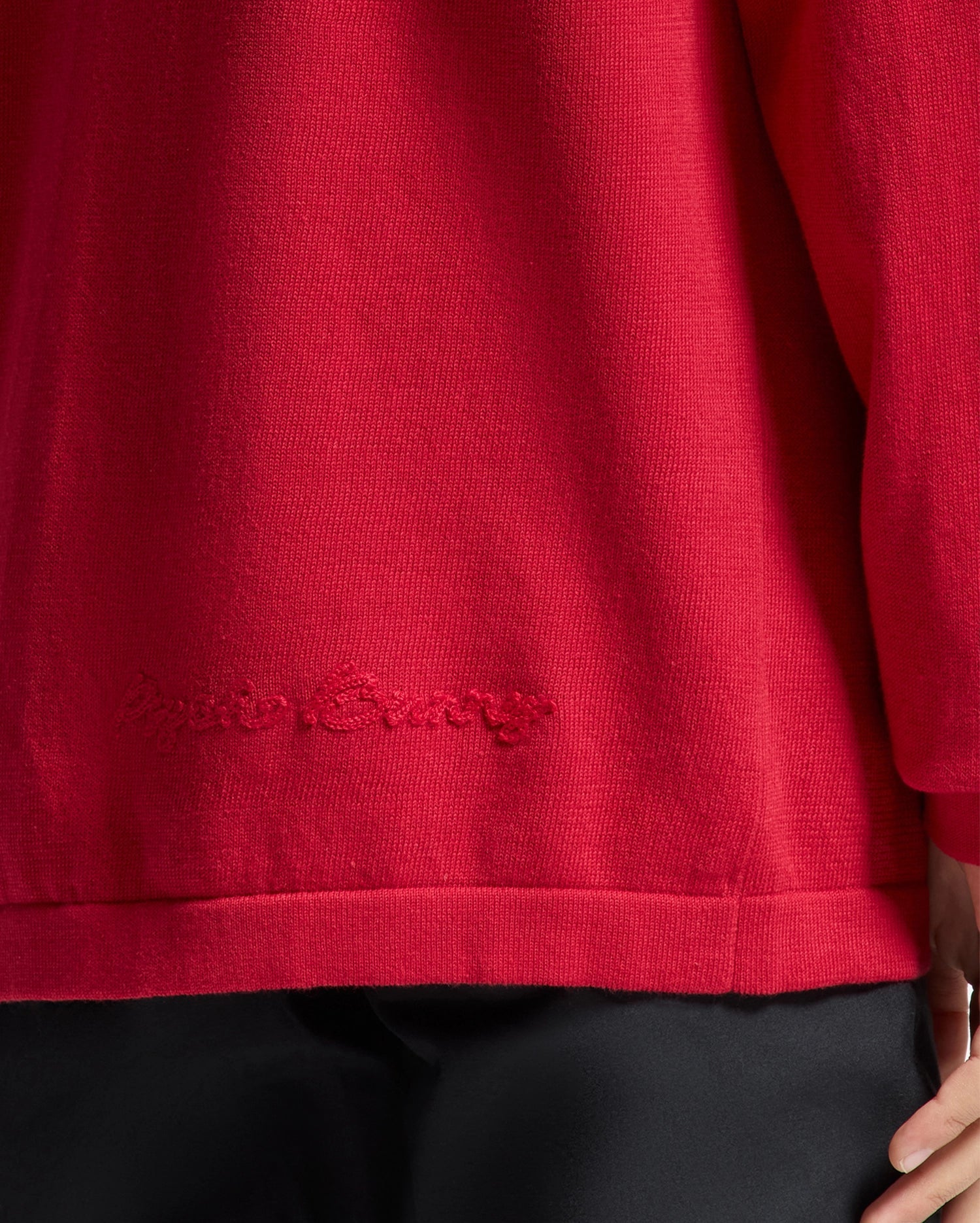 A person wearing a red KIDS COORY CHAINSTITCH COTTON SWEATER by Psycho Bunny, featuring "Gucci Camp" subtly chainstitched on the lower back. Made from extra fine cotton, it pairs perfectly with black pants. Only the lower back and part of the arm are visible.