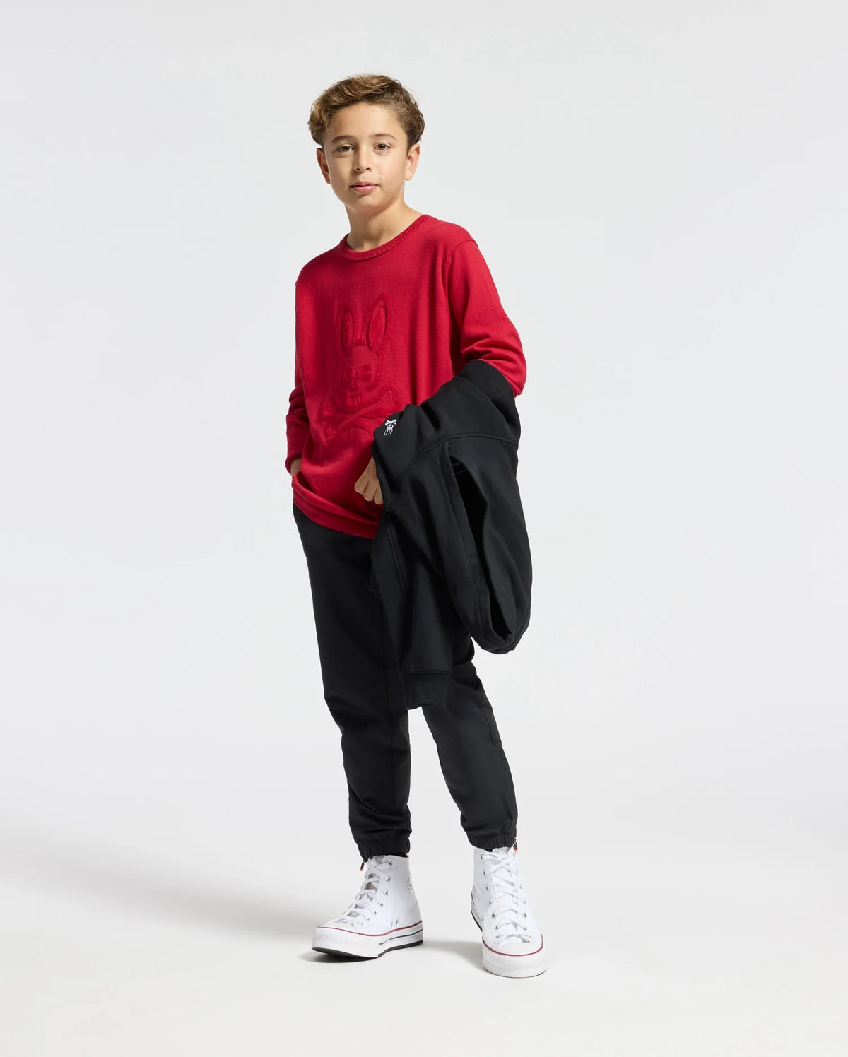 A young person stands confidently in Psycho Bunny's KIDS COORY CHAINSTITCH COTTON SWEATER, paired with black pants and white high-top sneakers, holding a black jacket over their arm against a plain white background.