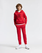 A person with curly hair is dressed in a red KIDS PIERCE ESSENTIAL FRENCH TERRY HOODIE by Psycho Bunny, paired with matching red sweatpants featuring a minimalistic graphic. They stand upright against a plain white background, wearing high-top white sneakers.
