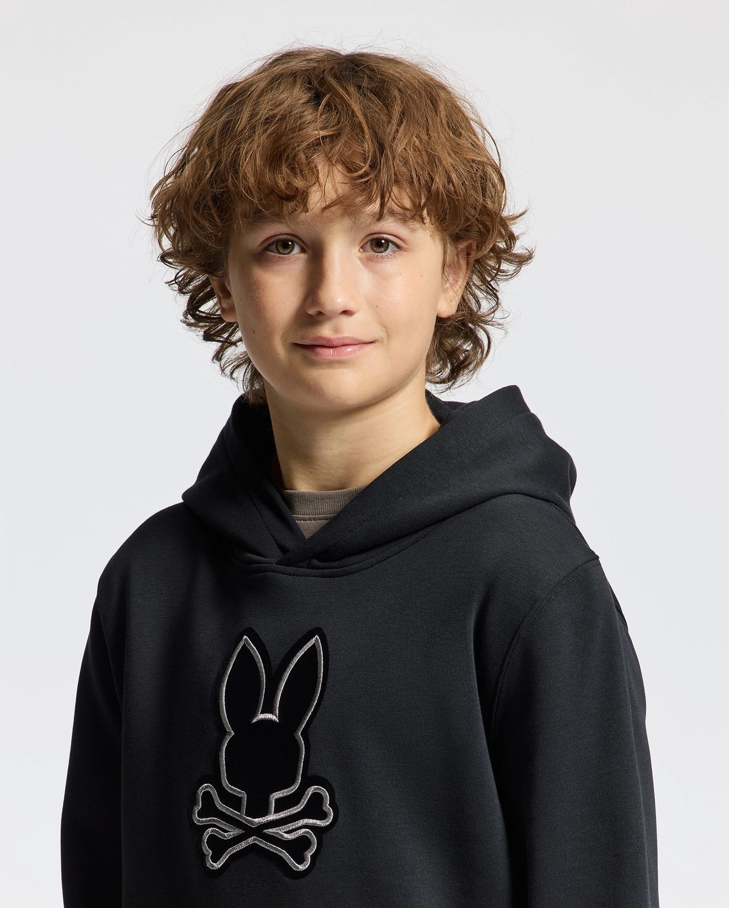 A young boy with curly brown hair is wearing a Psycho Bunny Kids Alexander Hoodie (B0H154E200) in black, featuring a bunny and crossbones design. This versatile pullover hoodie offers a comfortable relaxed fit, making it perfect for everyday adventures. He stands confidently against a plain white background.