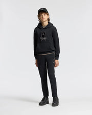 A person with curly hair stands facing forward, wearing the Psycho Bunny KIDS ALEXANDER HOODIE - B0H154E200 in black. This versatile pullover hoodie, featuring a bunny design and a comfortable relaxed fit, is paired with black pants and a black cap, standing out against the plain white background.