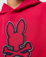 Close-up image of a person wearing a red KIDS WALTER LIGHTWEIGHT HOODIE - B0H476D200 by Psycho Bunny, featuring a stitched design of a bunny's head with crossed bones underneath. The design is predominantly black with red accents, and the surrounding stitching is white. This piece adds a modern twist to casual wear. The person's face is not visible.