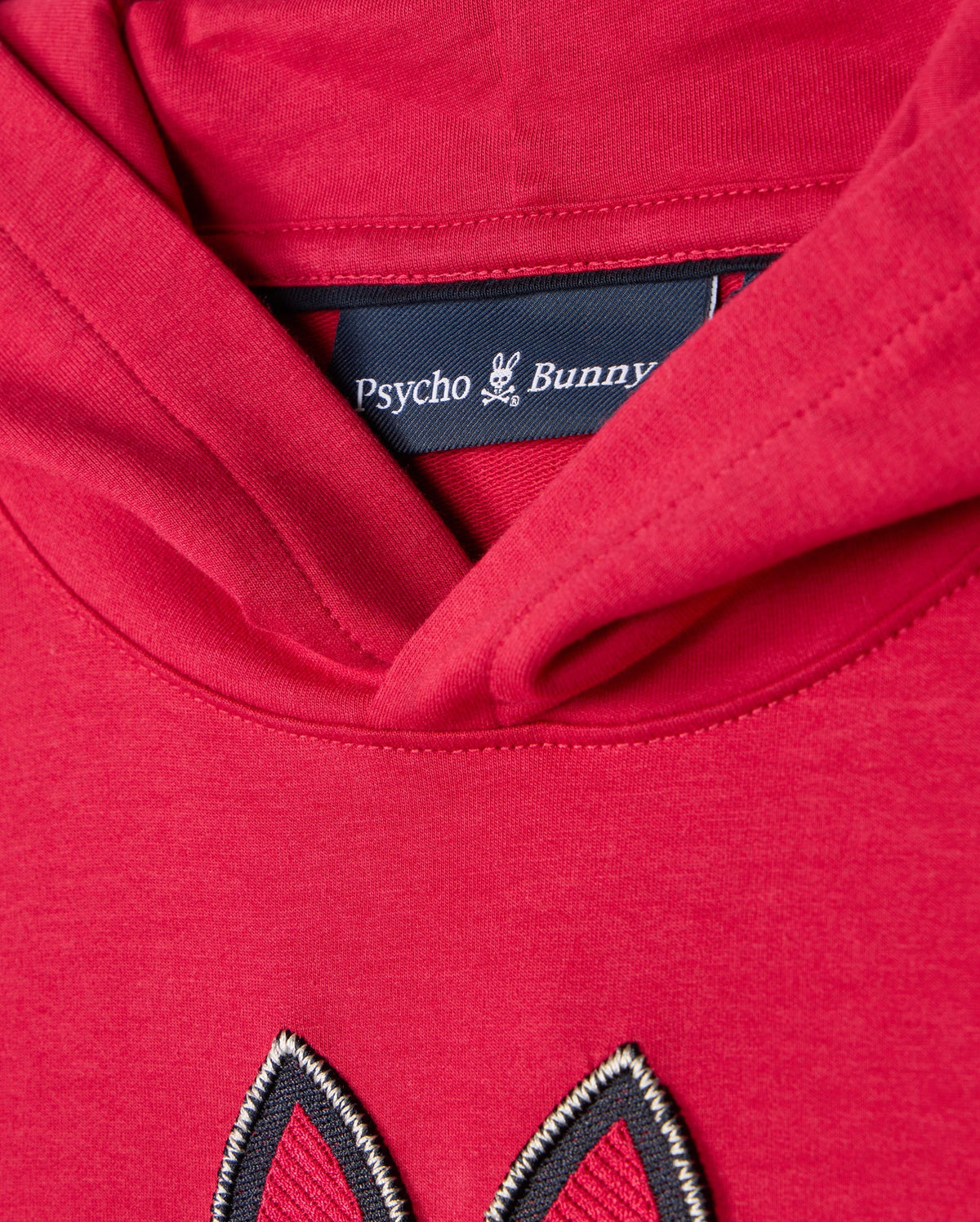 Close-up of the **KIDS WALTER LIGHTWEIGHT HOODIE - B0H476D200**, showcasing a red hoodie with a black clothing label inside the neckline, branded "Psycho Bunny" along with a small bunny logo. The hood is folded down, revealing a stitched design in matching red fabric below the label, adding a modern twist to this Micro French terry masterpiece.