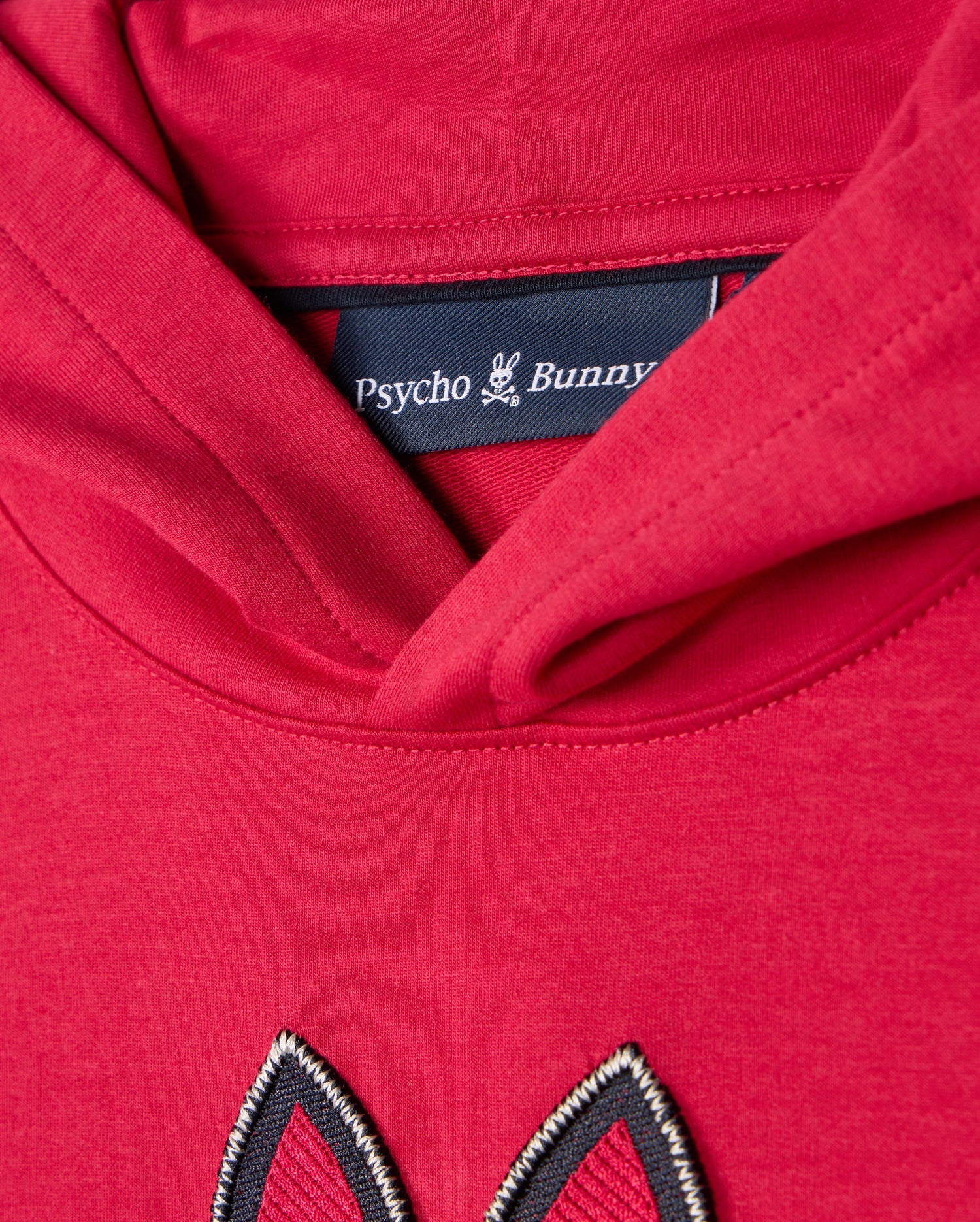 Close-up of the **KIDS WALTER LIGHTWEIGHT HOODIE - B0H476D200**, showcasing a red hoodie with a black clothing label inside the neckline, branded "Psycho Bunny" along with a small bunny logo. The hood is folded down, revealing a stitched design in matching red fabric below the label, adding a modern twist to this Micro French terry masterpiece.
