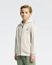 A young boy with short blond hair smiles slightly, wearing a light beige KIDS LEVY ESSENTIAL ZIP FLEECE HOODIE from Psycho Bunny, featuring a small embroidered logo on the chest, paired with green pants. Crafted from soft cotton for kids' comfort against a simple background.