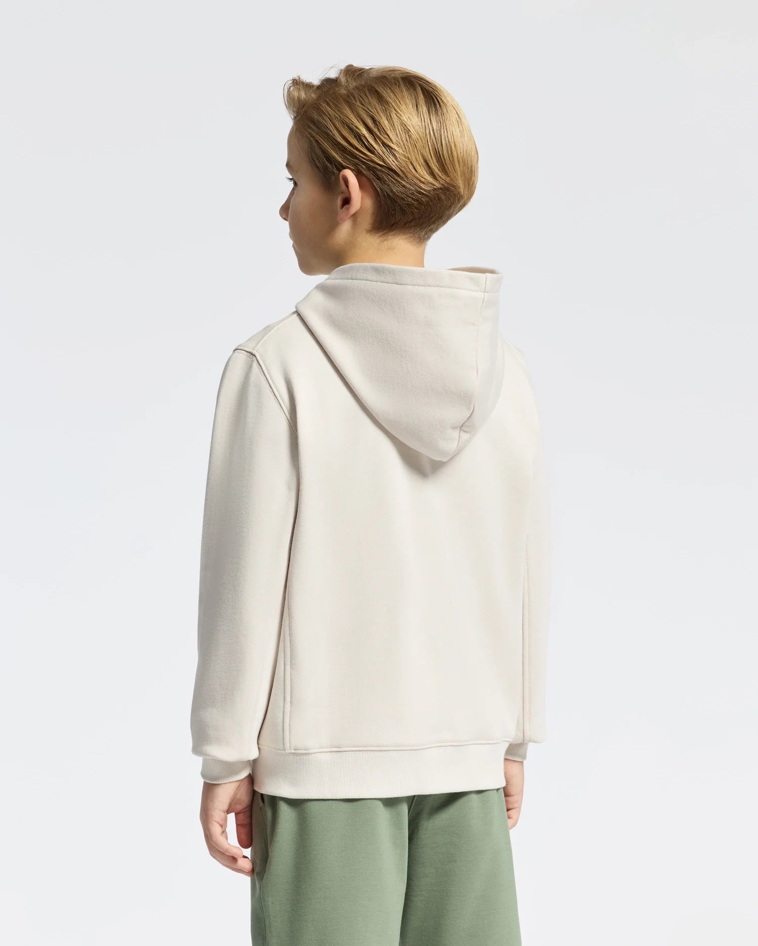A child with short blond hair, wearing a cream Psycho Bunny KIDS LEVY ESSENTIAL ZIP FLEECE HOODIE - B0H720D200 and green pants, stands facing away against a plain white background. This high-quality cotton hoodie is perfect for those seeking stylish kids' clothing.