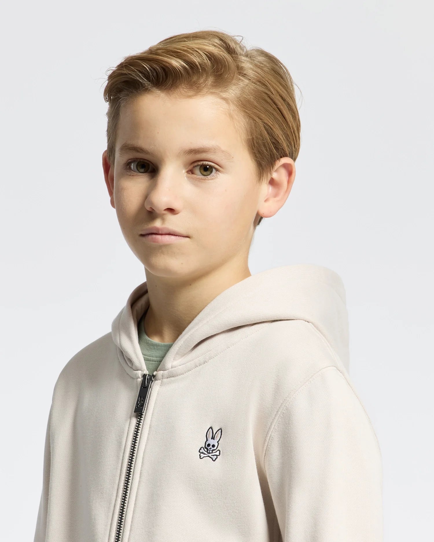 A young boy with short blond hair wears a Psycho Bunny Kids Levy Essential Zip Fleece Hoodie (B0H720D200), a light-colored design featuring a small rabbit logo on the chest. The plain, light gray background accentuates this cotton kids' clothing staple.