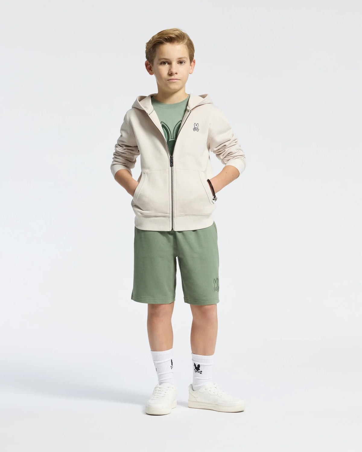 A boy stands confidently in a Psycho Bunny KIDS LEVY ESSENTIAL ZIP FLEECE HOODIE - B0H720D200, crafted from soft French terry. The light zip-up hoodie complements his green shirt and matching shorts. With hands in pockets, he sports white sneakers and socks featuring a graphic, set against a plain white background.