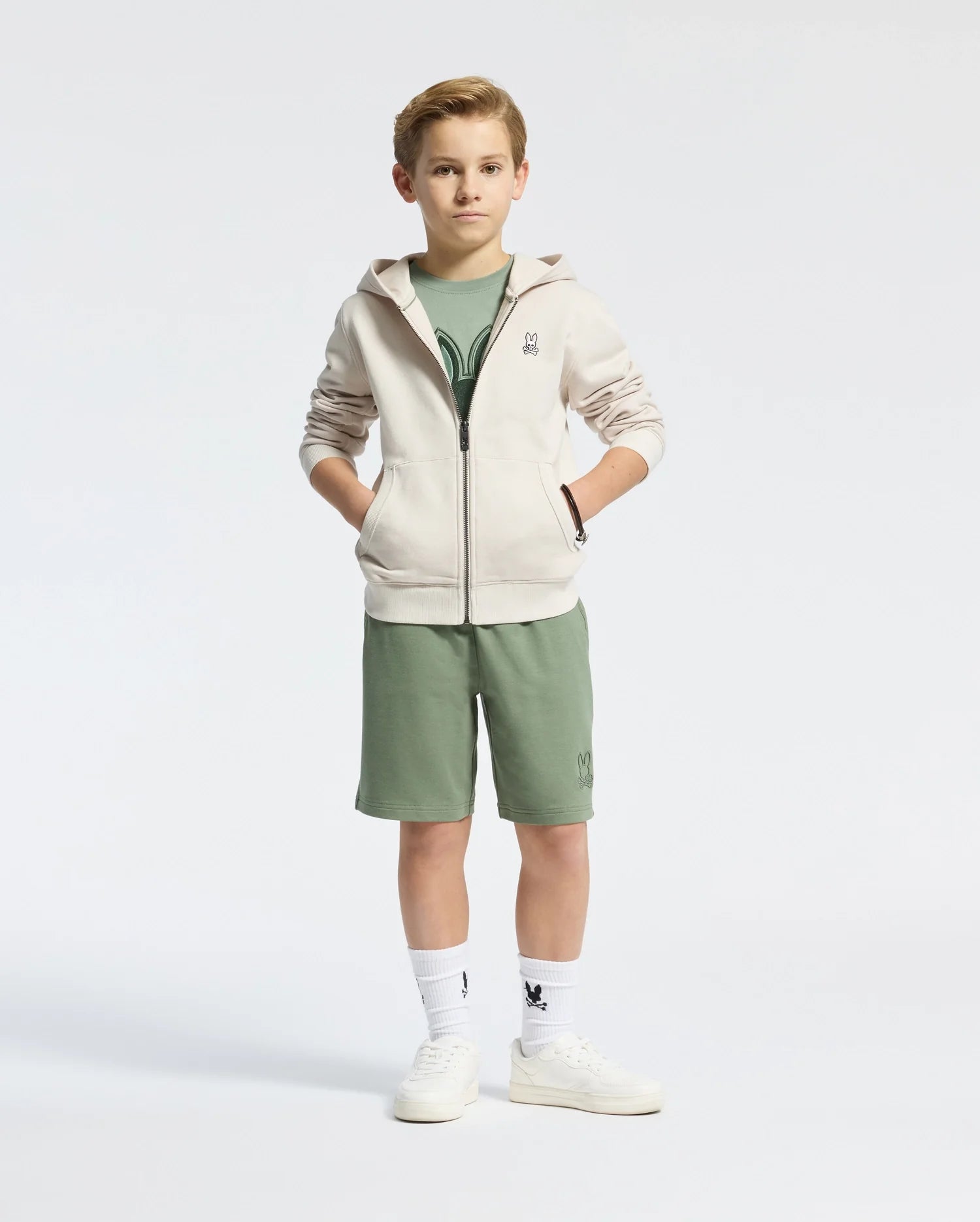 A boy stands confidently in a Psycho Bunny KIDS LEVY ESSENTIAL ZIP FLEECE HOODIE - B0H720D200, crafted from soft French terry. The light zip-up hoodie complements his green shirt and matching shorts. With hands in pockets, he sports white sneakers and socks featuring a graphic, set against a plain white background.