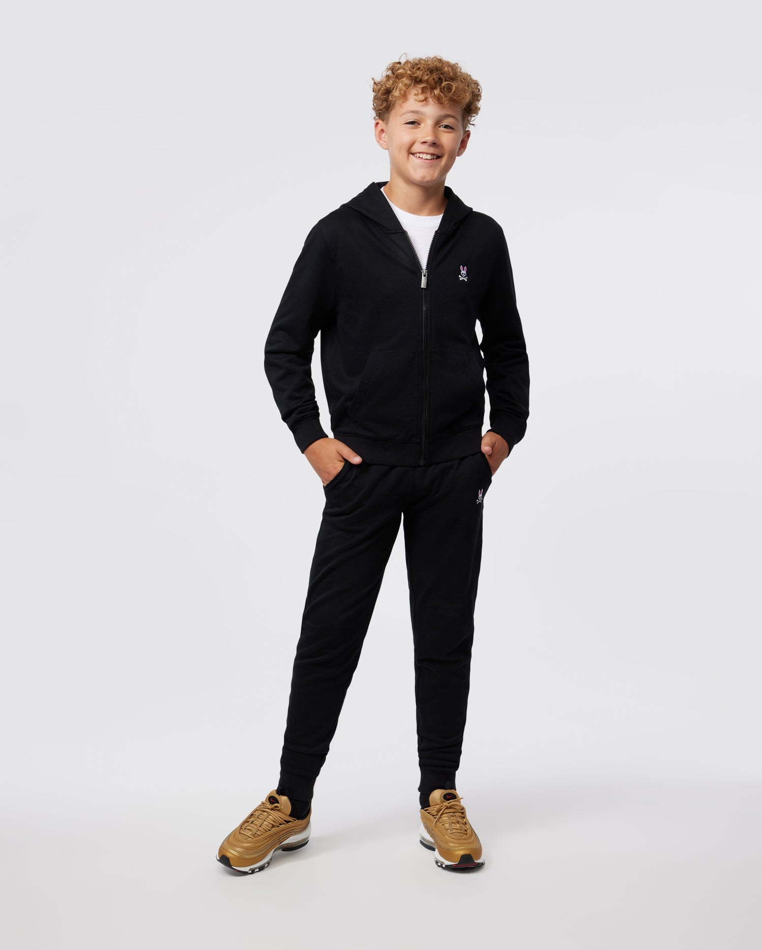 A smiling young boy with curly hair, dressed in a black Psycho Bunny KIDS CLASSIC FRENCH TERRY ZIP HOODIE - B0H826ARFT with a kangaroo pocket and matching pants, stands confidently with hands in pockets against a white background. He wears brown