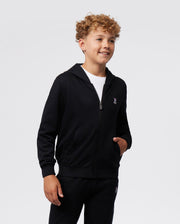 A smiling young boy with curly hair wearing a black Psycho Bunny KIDS CLASSIC FRENCH TERRY ZIP HOODIE - B0H826ARFT and matching pants, standing against a plain white background. He has his hand in a kangaroo pocket.