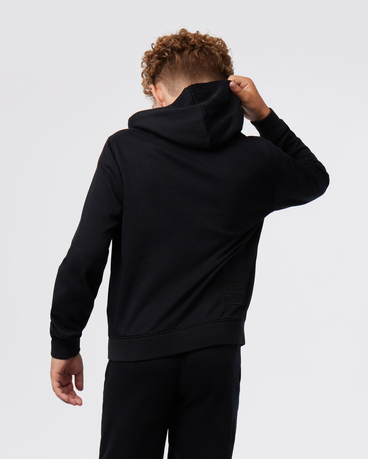A young person seen from behind wearing a black Psycho Bunny KIDS CLASSIC FRENCH TERRY ZIP HOODIE and matching pants, adjusting the hood with one hand. The hoodie features a subtle square pattern on the lower right side.