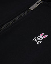 Close-up of a black Psycho Bunny KIDS CLASSIC FRENCH TERRY ZIP HOODIE with an embroidered bunny logo featuring a skull with crossed bones and pink ears, next to a partially zipped silver zipper.