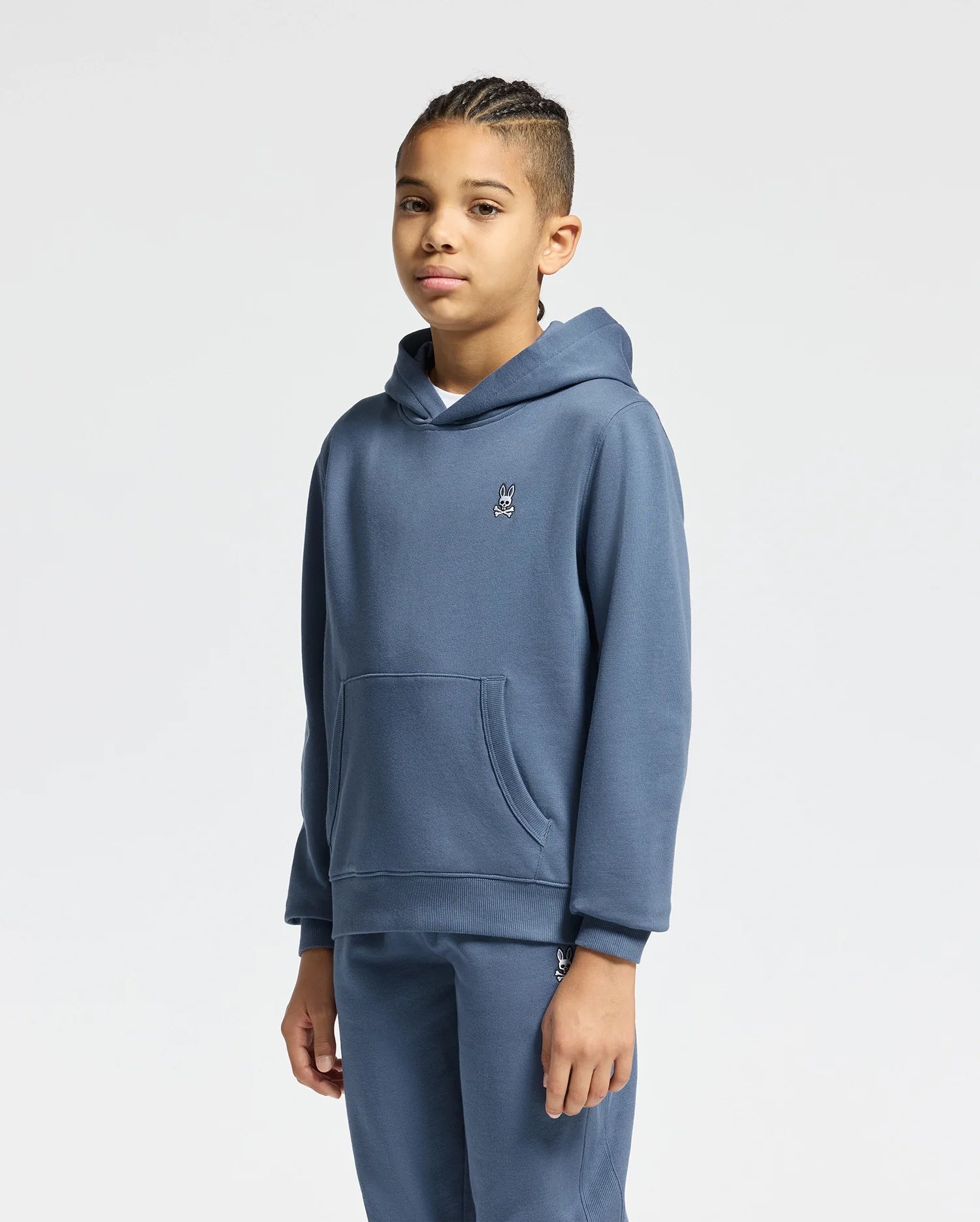 A child with braided hair is wearing a KIDS LEVY ESSENTIAL FLEECE HOODIE from Psycho Bunny, crafted entirely from cotton. Paired with matching fleece sweatpants in blue, both featuring a discreet embroidered logo on the chest, this ensemble stands out against the simple white background for a cozy and stylish look.