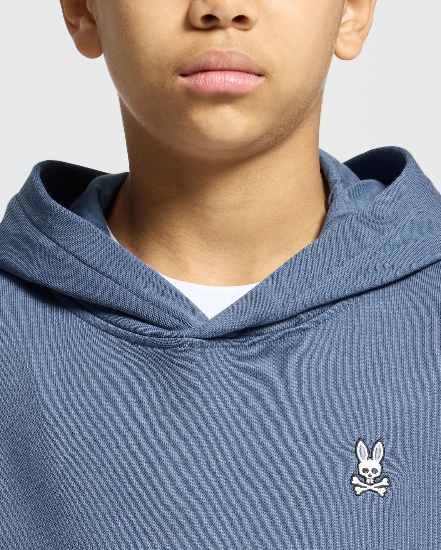 A person is wearing a blue Psycho Bunny Kids Levy Essential Fleece Hoodie (B0H859D200), featuring a small white bunny and crossbones logo on the chest. This cozy 100% cotton hoodie, layered over a white shirt, provides comfort while partially obscuring the person's face.